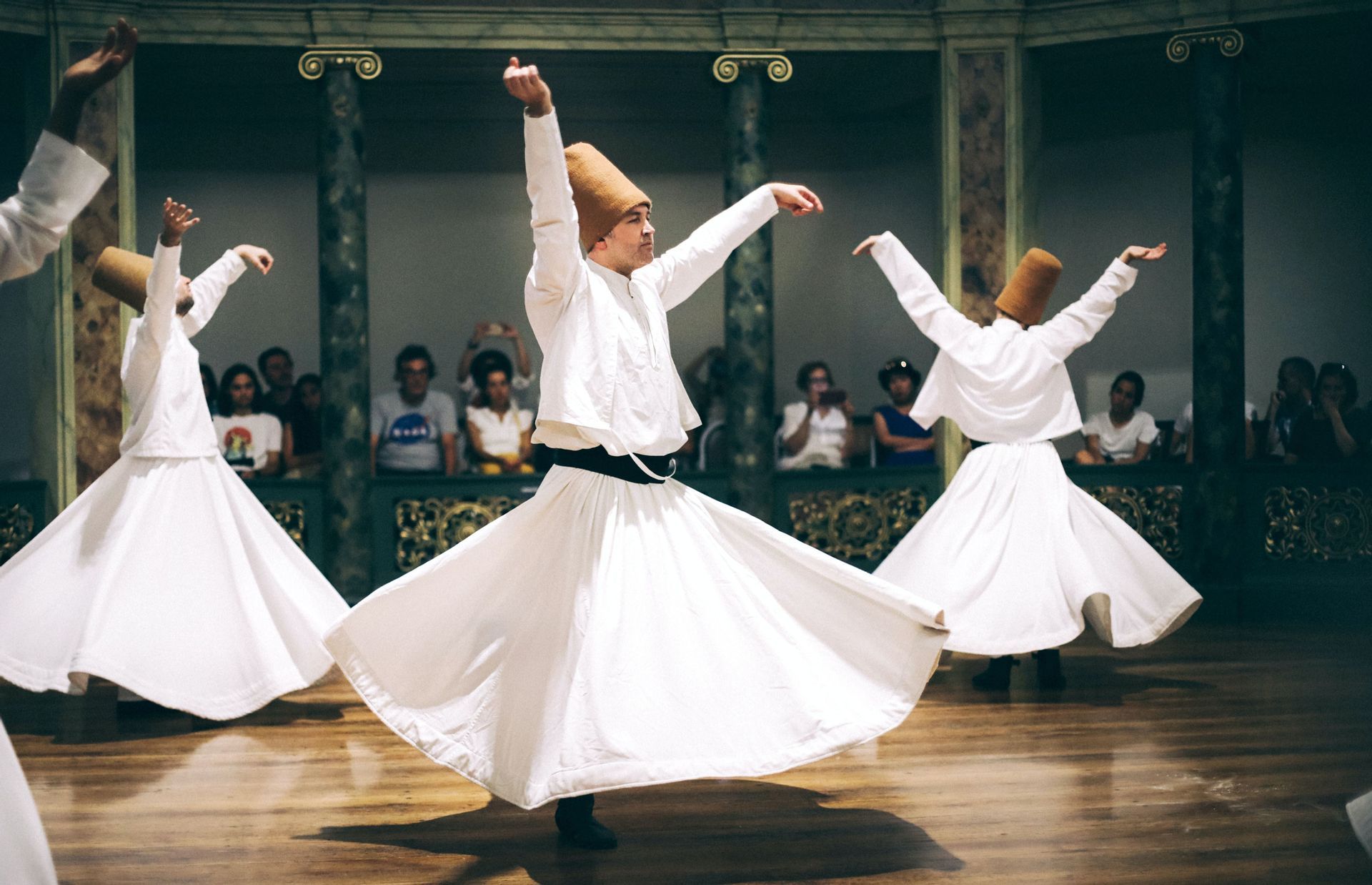 Several men in white robes and tall hats perform the Whirling Dervish dance on a wooden floor as an audience watches.