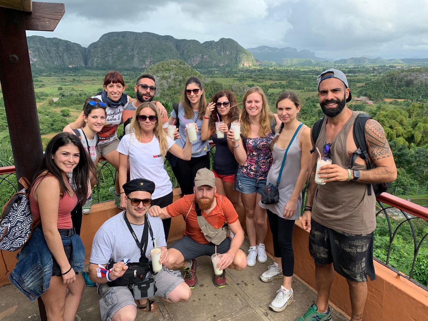 Un gruppo WeRoad in posa per una foto con bevande su un punto panoramico che si affaccia su una lussureggiante valle verde e le montagne.