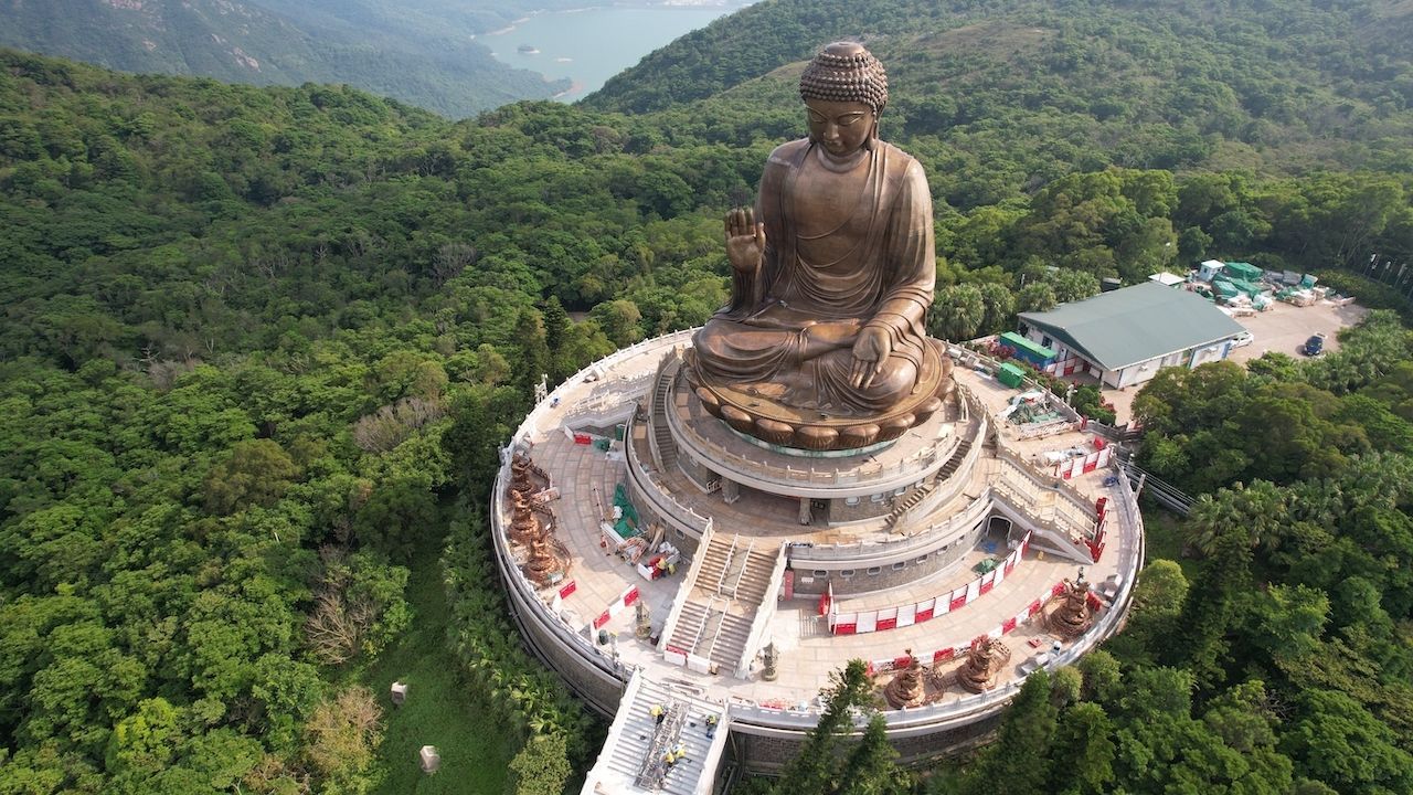 Veduta aerea di una grande statua di Buddha in bronzo, situata in cima a una piattaforma a più livelli su una montagna coperta da una fitta foresta verde.