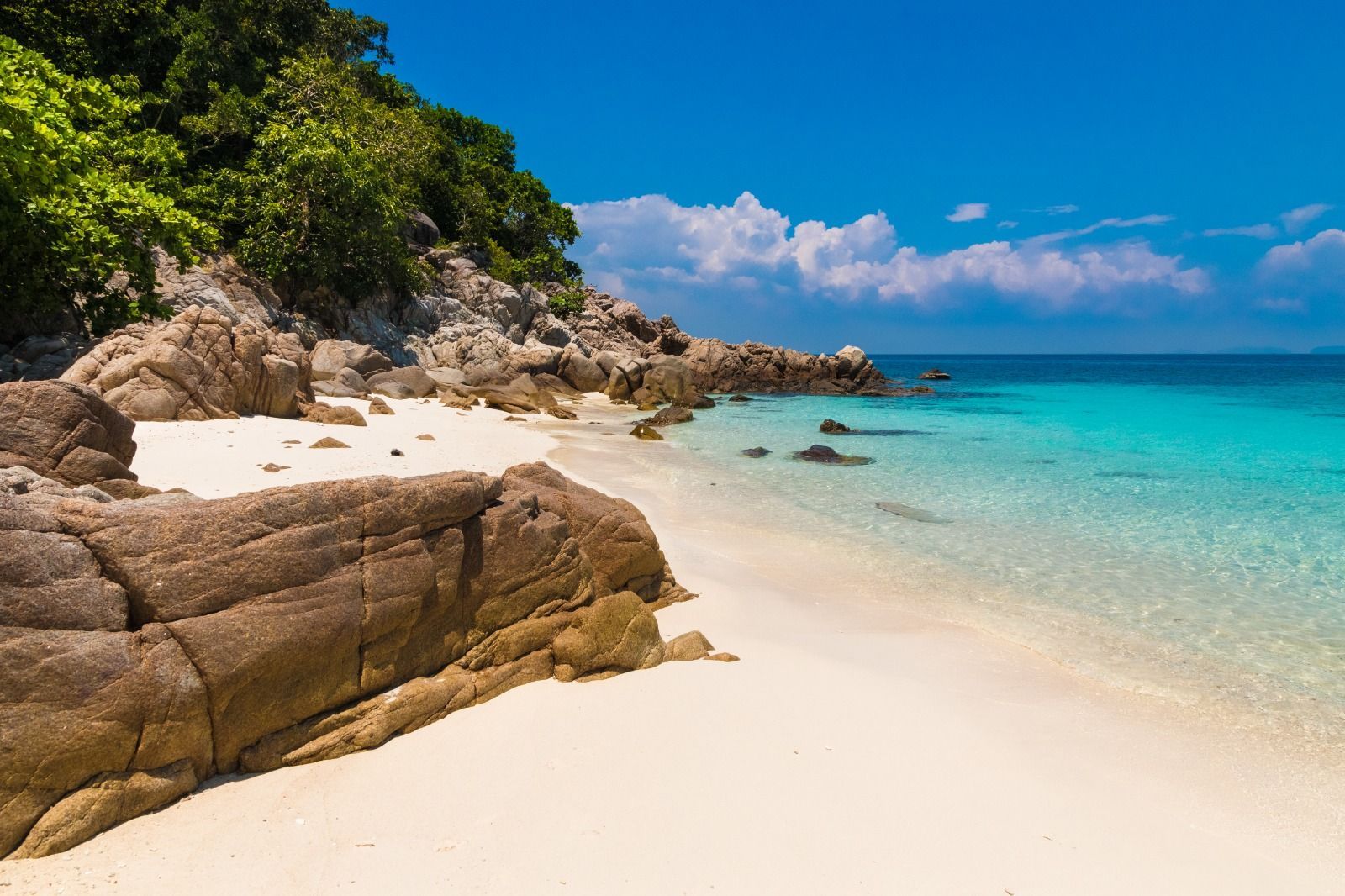 Una playa aislada de arena blanca con grandes rocas, rodeada de exuberante vegetación y aguas tranquilas y turquesas bajo un cielo azul.