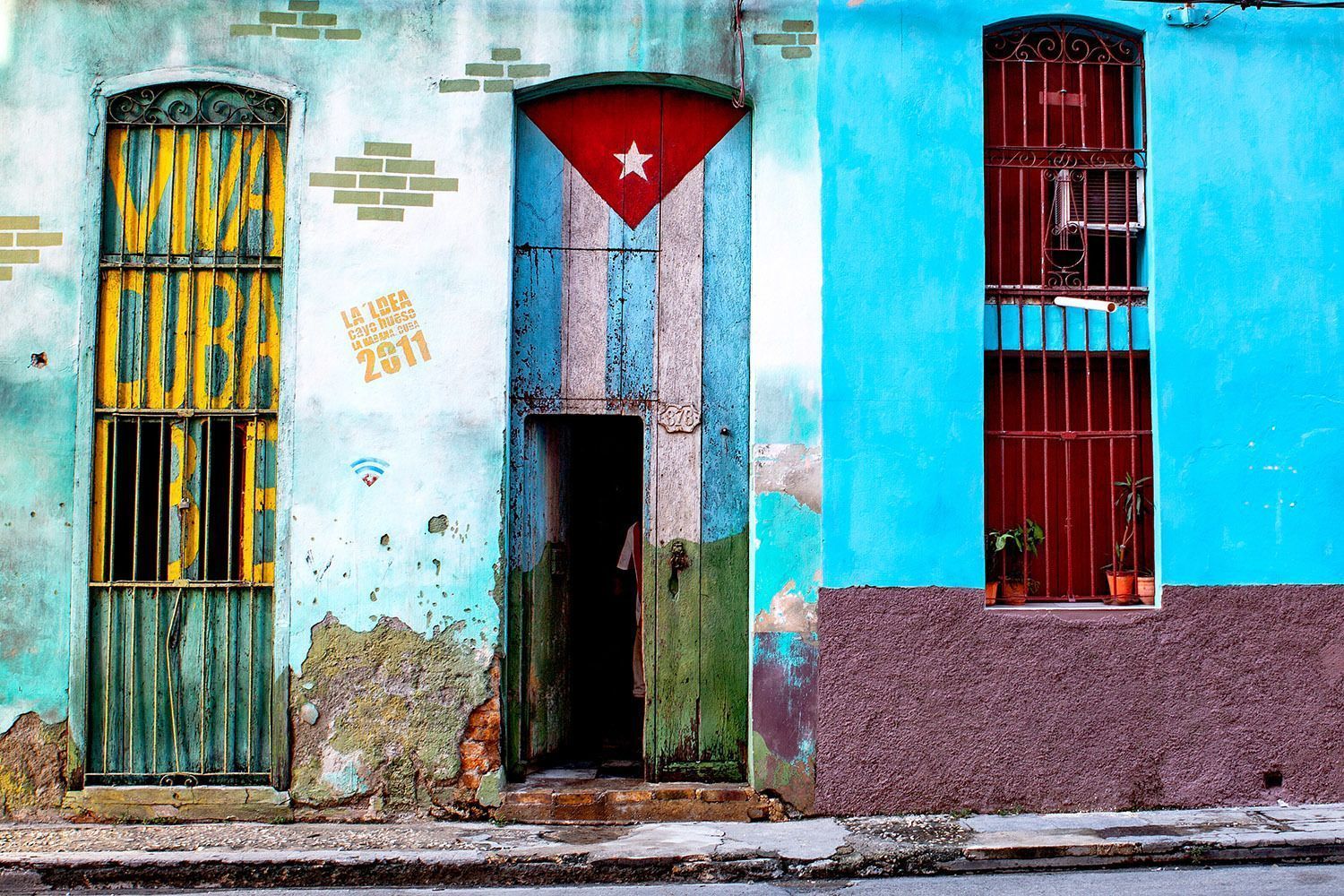 Una pared colorida y desgastada con una puerta pintada con la bandera cubana, una puerta enrejada verde y amarilla, y una ventana enrejada roja.