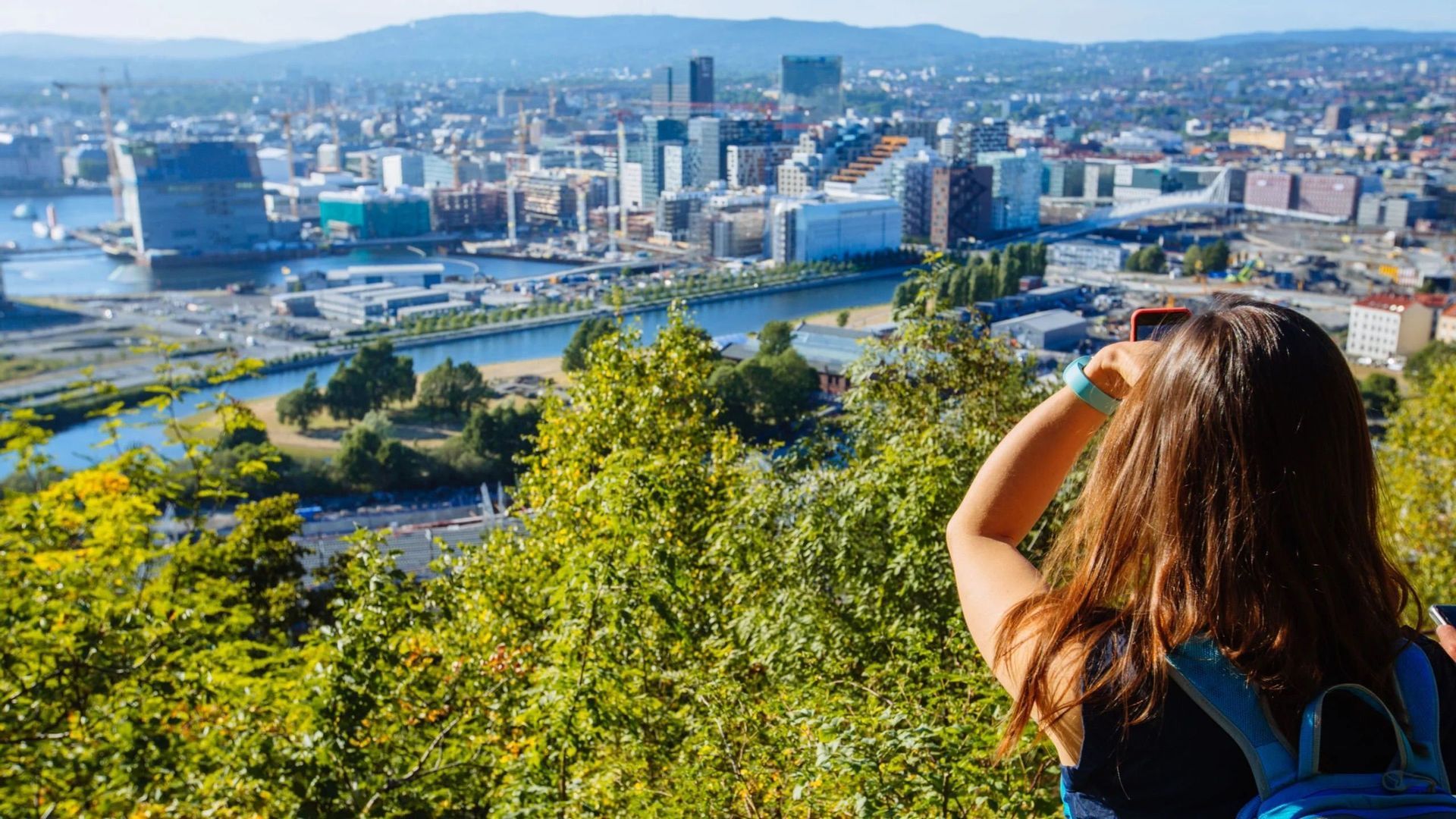 Una donna con uno zaino, vista da dietro, fotografa un moderno paesaggio urbano con un fiume da un punto di vista elevato.
