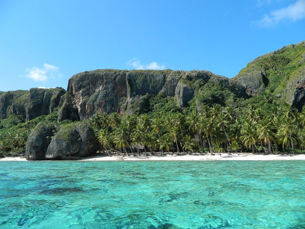 Acqua cristallina turchese di fronte a una spiaggia di sabbia bianca fiancheggiata da palme, con una grande scogliera ricoperta di verde sullo sfondo.