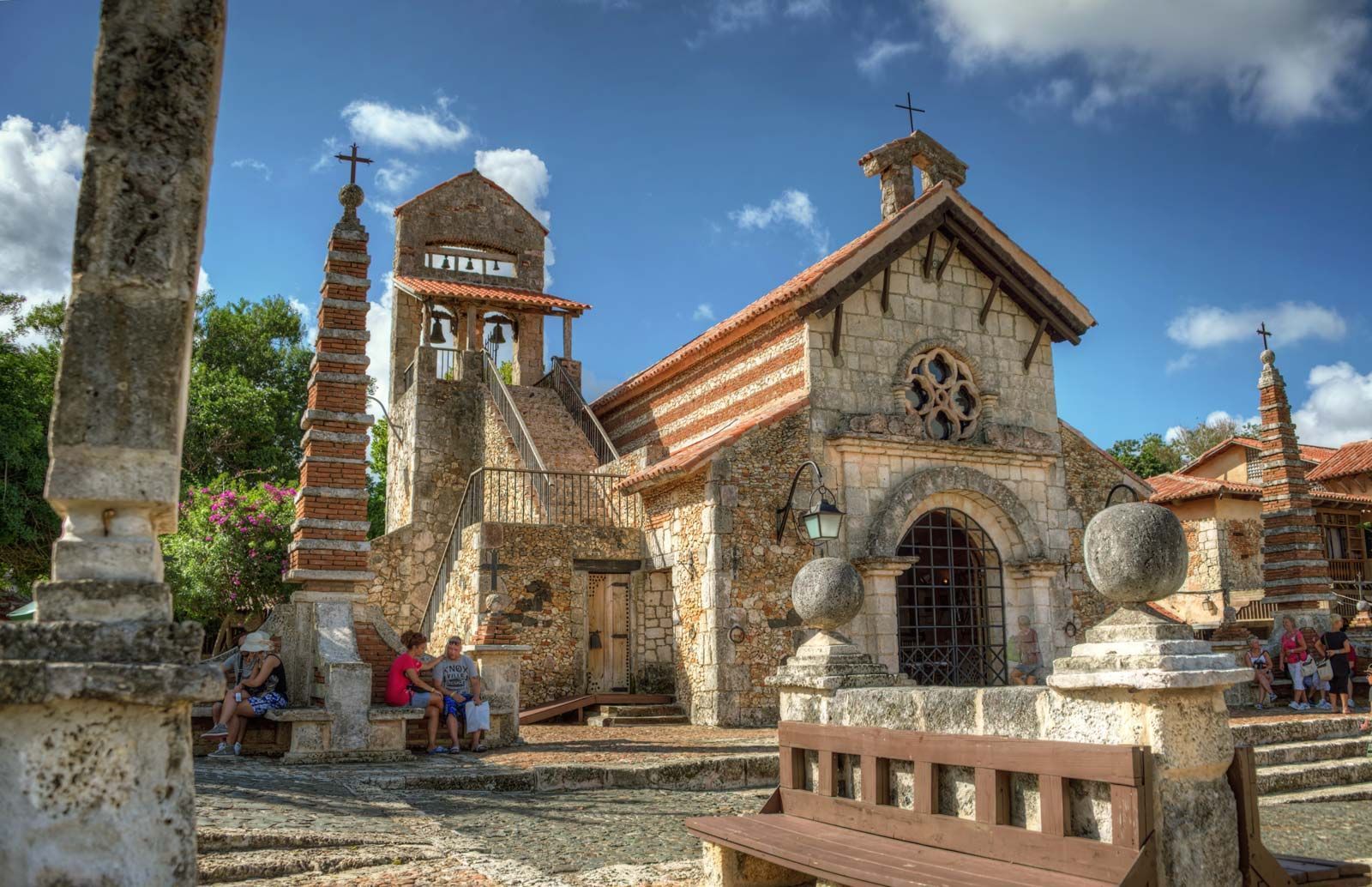Una chiesa rustica in pietra con campanile e un obelisco in una piazza soleggiata con persone che riposano sulle panchine.
