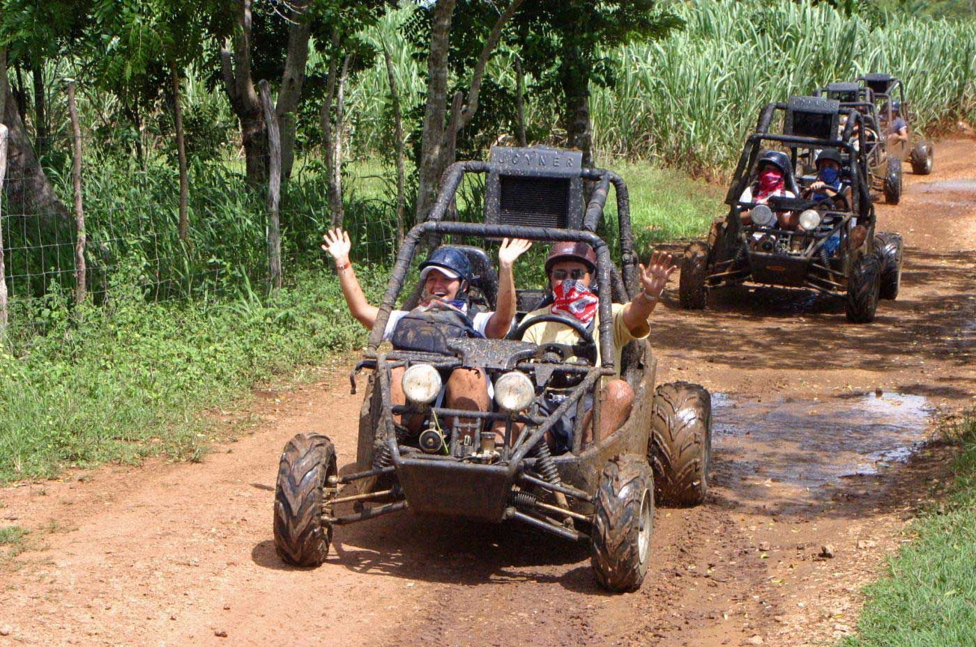 Due persone in un viaggio di gruppo WeRoad salutano dalla parte anteriore di un dune buggy fangoso, alla guida di un convoglio lungo un sentiero sterrato.