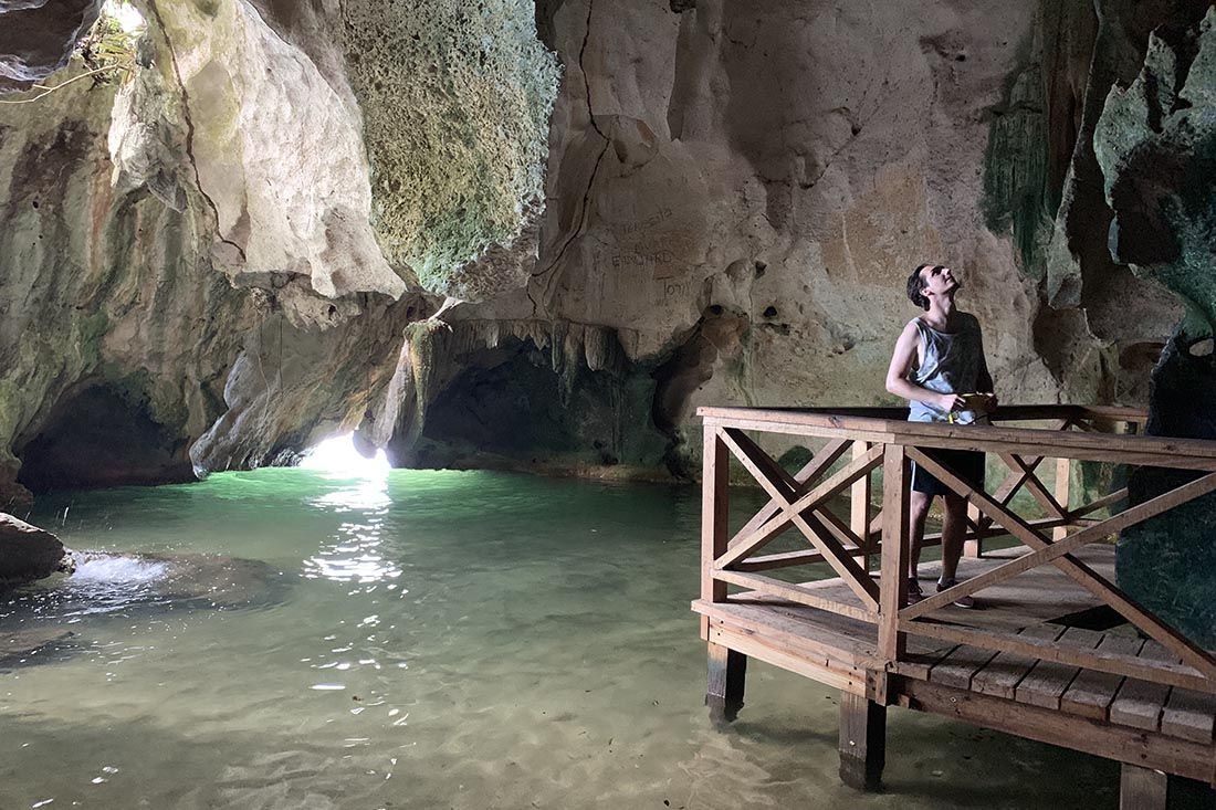 Una persona si trova su una piattaforma di legno, guardando in alto all'interno di un'ampia grotta con una pozza d'acqua verde illuminata da un'apertura.