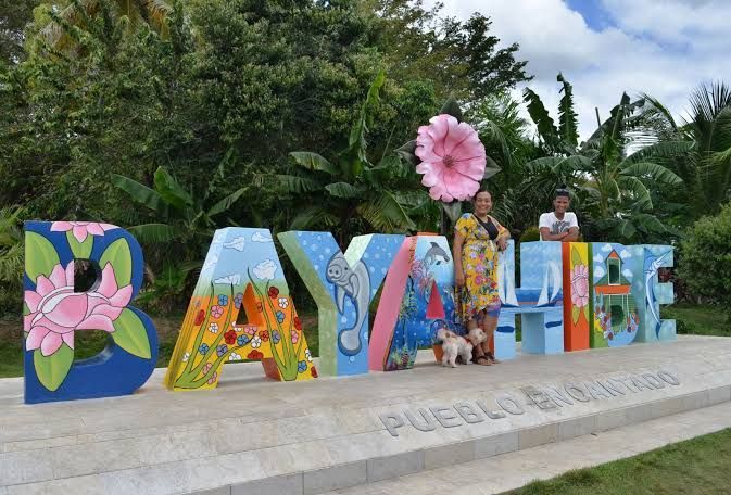 Una donna, un ragazzo e un cagnolino posano accanto a un grande cartello colorato con la scritta 'Bayahibe' all'aperto.