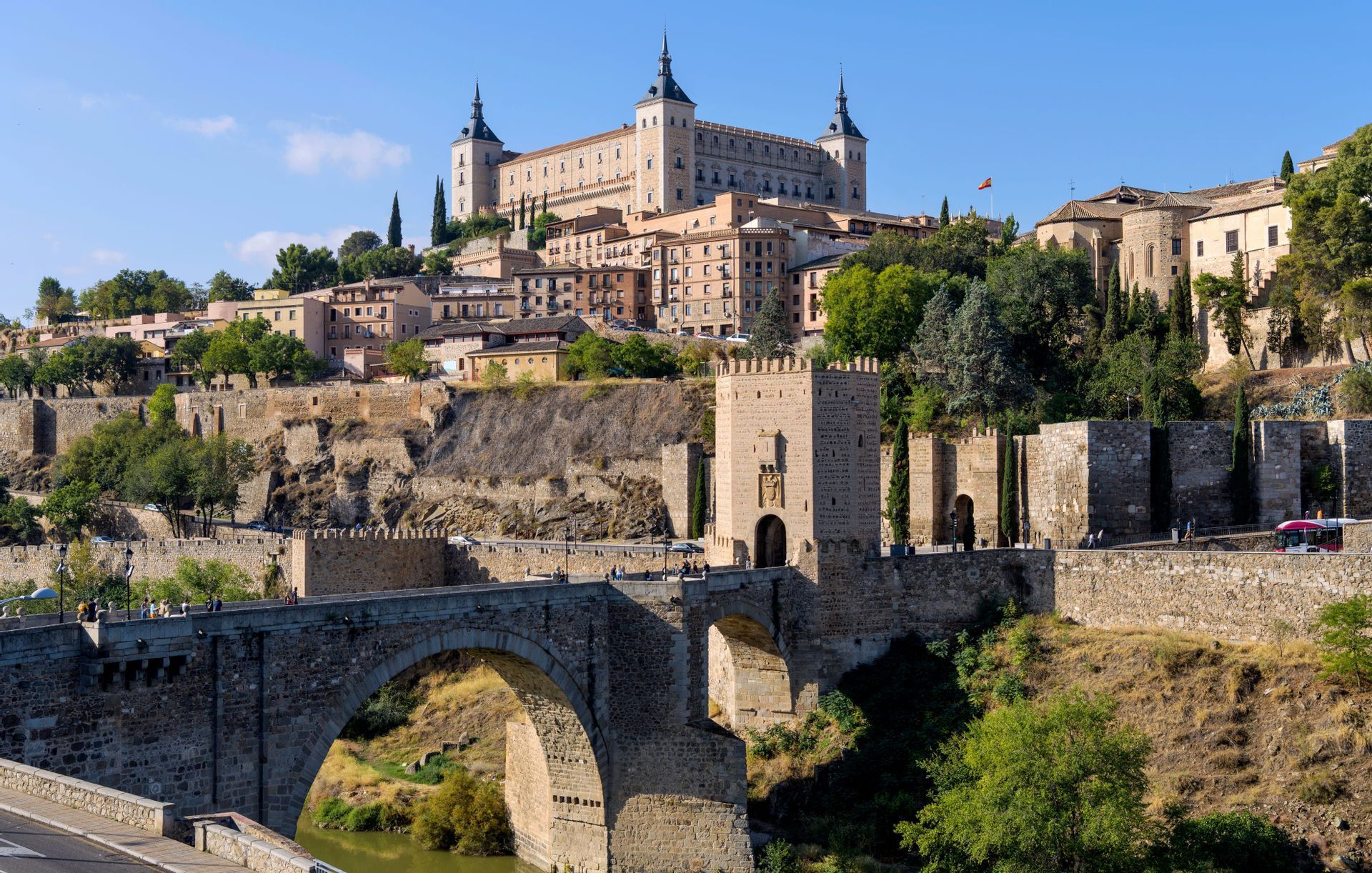 Un grande ponte in pietra con archi, che attraversa un fiume, porta a una storica città fortificata su una collina sotto un cielo azzurro e sereno.