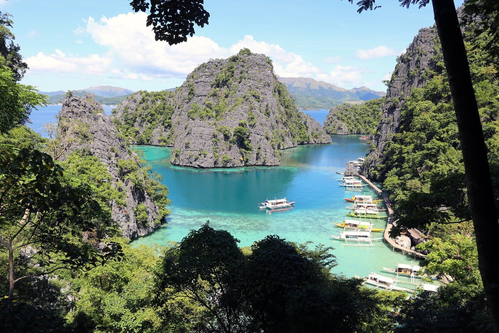 Una vista dall'alto di una laguna tropicale con acqua turchese, circondata da ripide scogliere ricoperte di giungla e piena di barche a bilanciere.