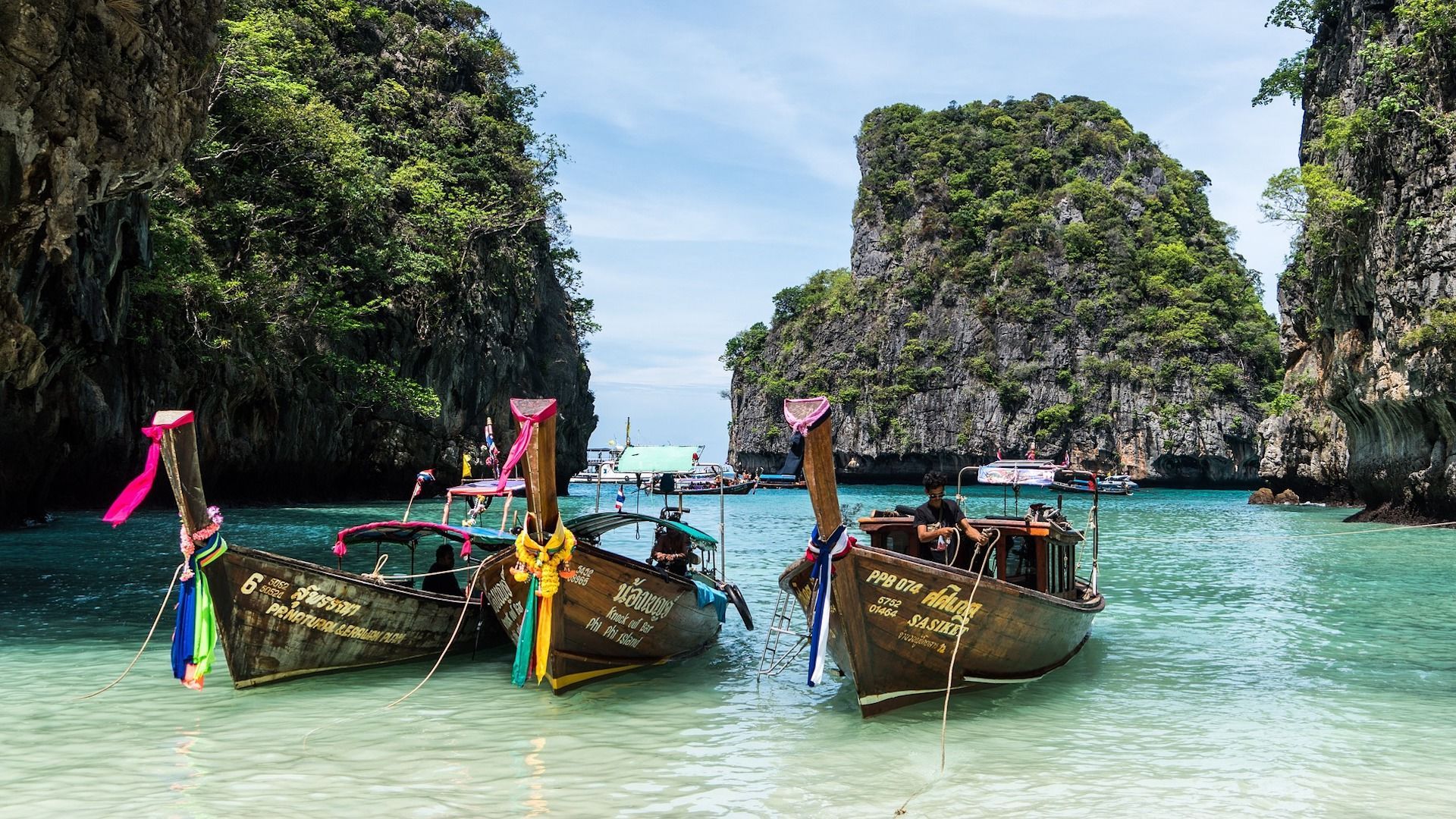 Barche tradizionali in legno a coda lunga, adornate con nastri colorati, ormeggiate in acque turchesi in una baia circondata da lussureggianti scogliere rocciose.
