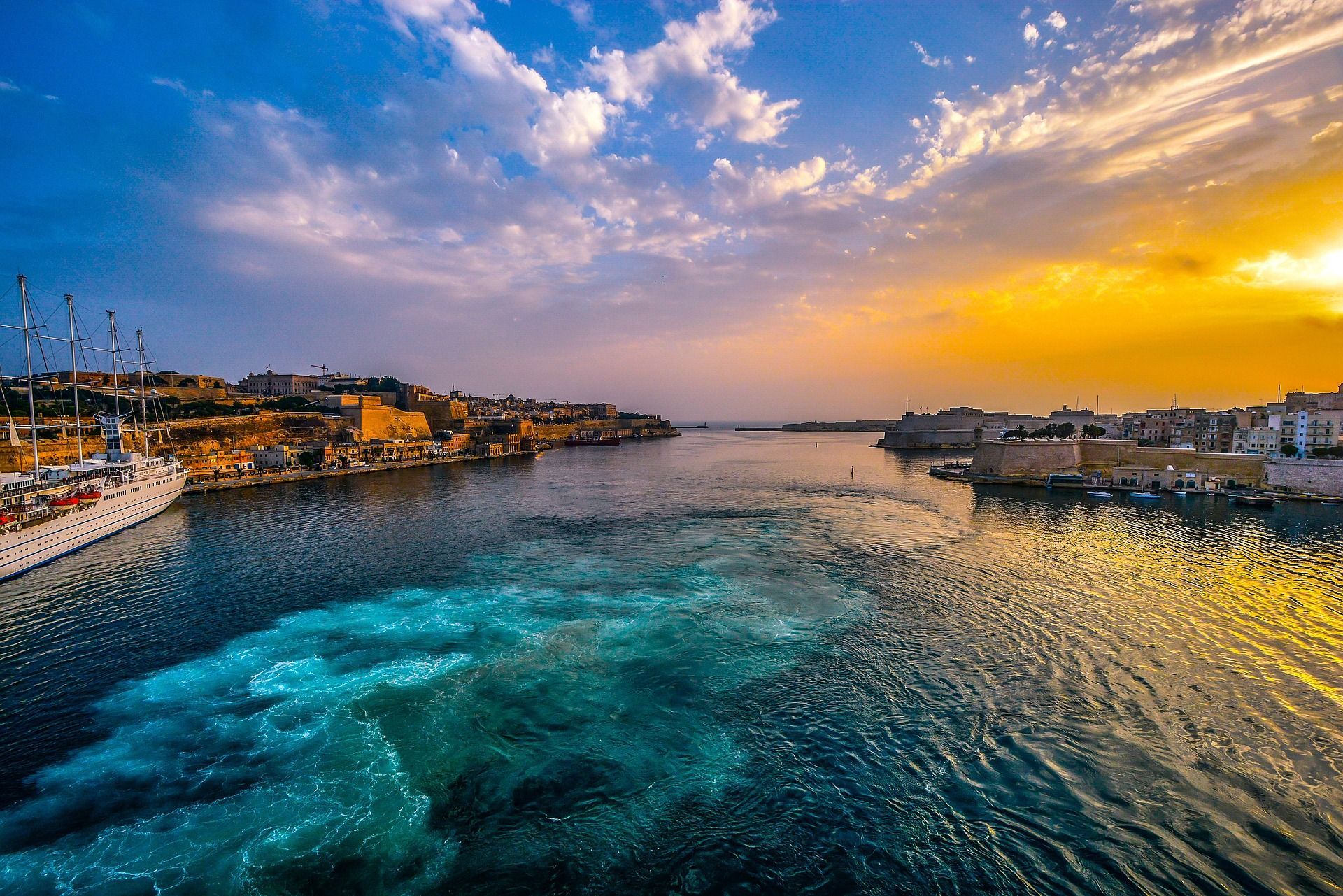 A view from the water of a historic city harbor at sunset, with a large docked ship and a turbulent wake in the foreground.