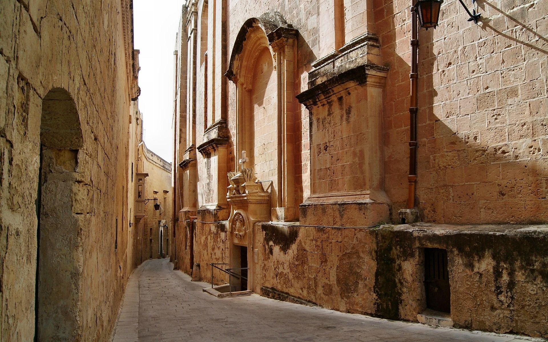A narrow cobblestone alleyway curves between tall, weathered stone buildings under bright sunlight.