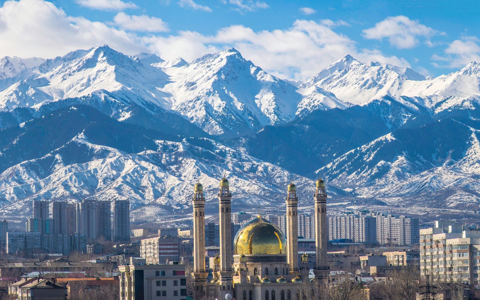 A mosque with a golden dome and four minarets stands in a cityscape at the foot of a large, snow-capped mountain range.