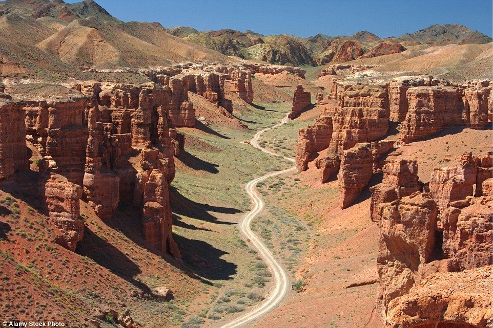 Vista aérea de un camino de tierra sinuoso que atraviesa un cañón profundo con formaciones rojas y vegetación verde.