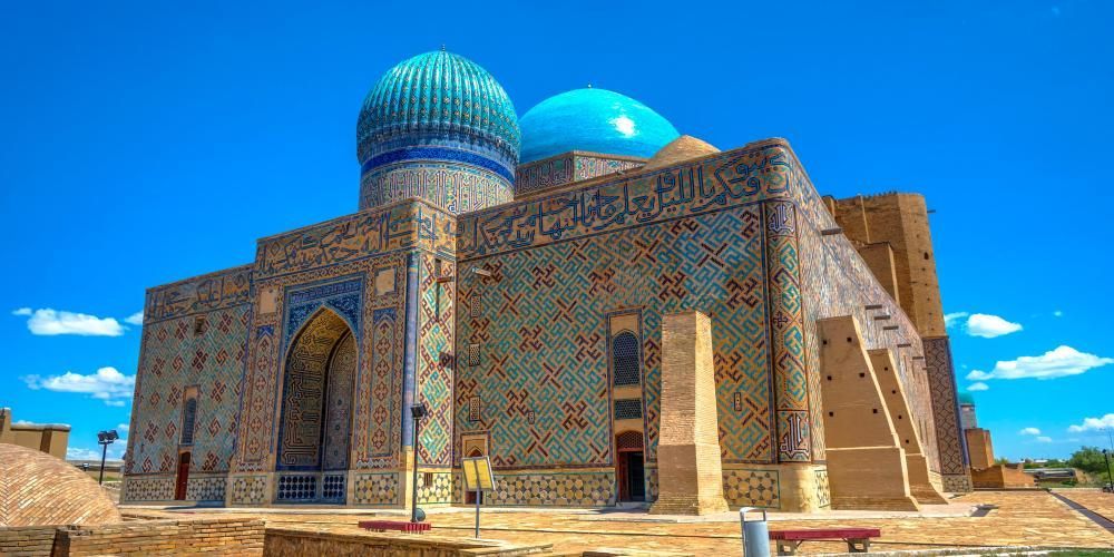 An ornate mausoleum with two blue domes and walls covered in intricate blue and green tile mosaics stands under a clear blue sky.