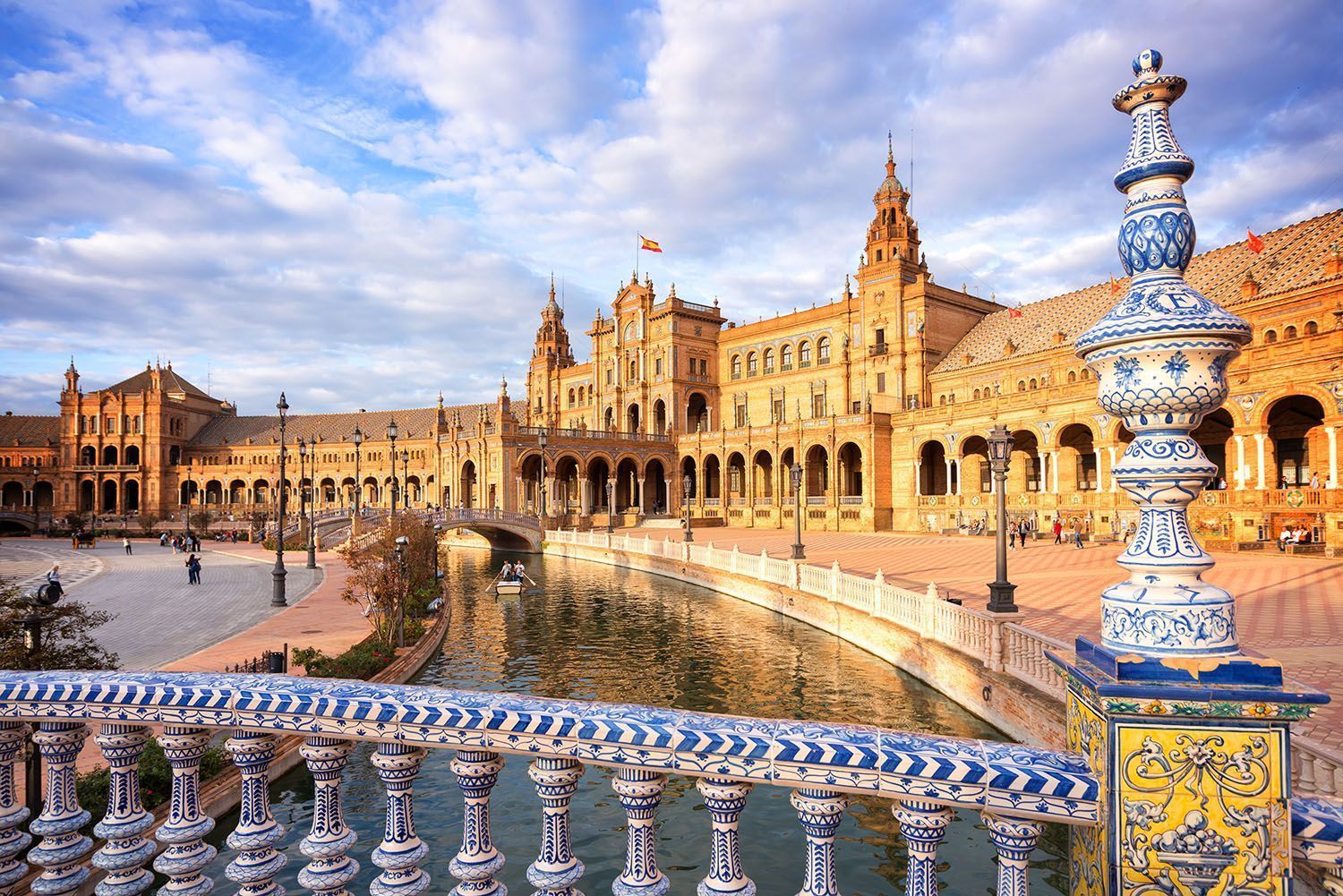 An ornate building with a canal is seen from a bridge with a decorative blue and white tiled balustrade under a partly cloudy sky.