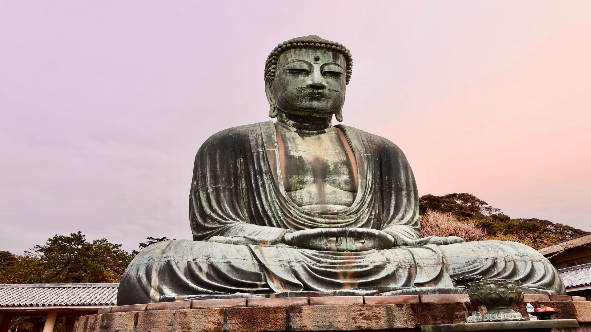 Una grande statua in bronzo patinato del Buddha si trova in posa meditativa su una base di pietra all'aperto, con alberi sullo sfondo.