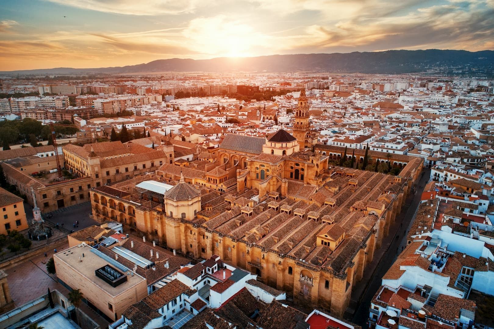 An aerial view of a sprawling city with terracotta roofs centered around a large historic cathedral complex at sunset.