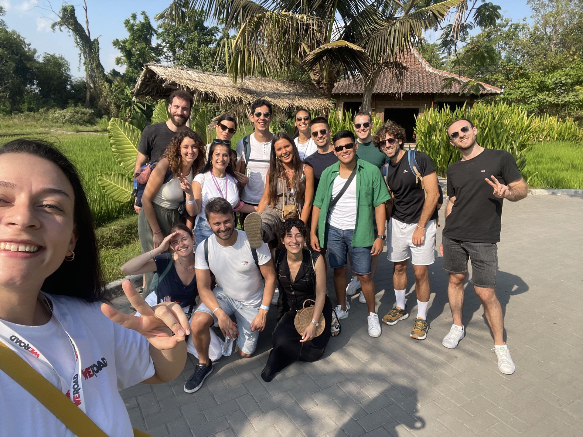 Un grupo de WeRoad posa para una selfie sonriente al aire libre, con palmeras y una cabaña tradicional de fondo.
