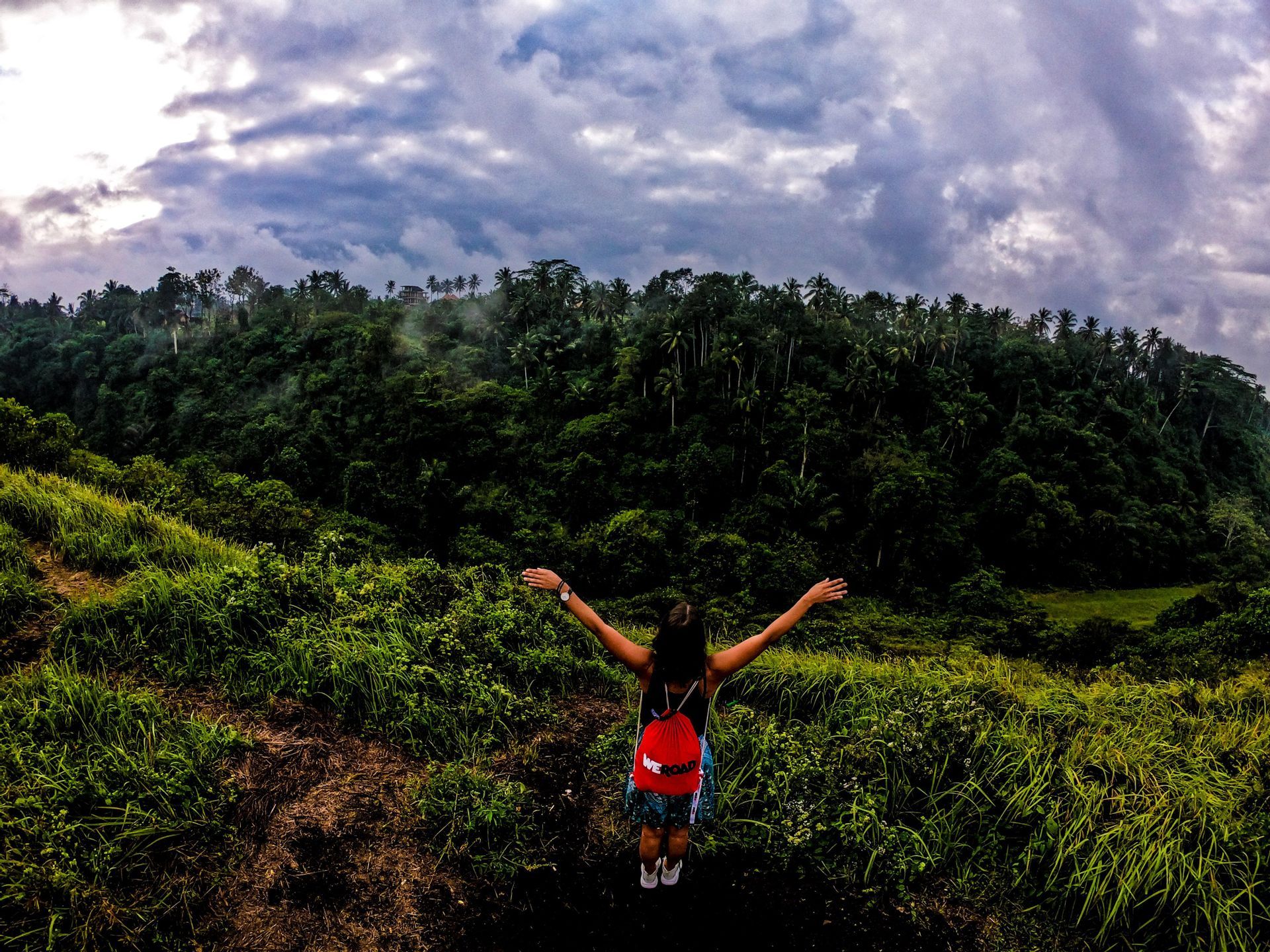 Una mujer con una mochila WeRoad roja, con los brazos extendidos en una colina de hierba, con vistas a un denso bosque verde bajo un cielo nublado.