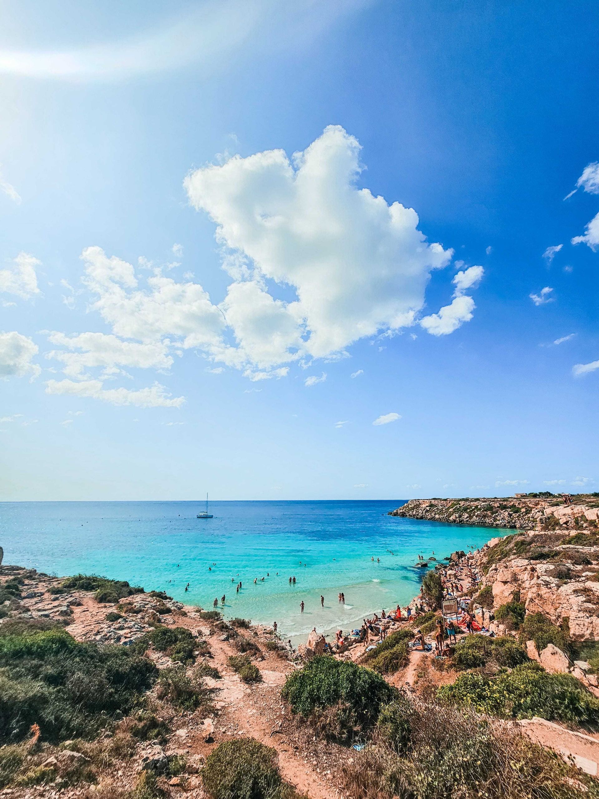 Una veduta dall'alto di una cala con acqua turchese splendente, delimitata da scogliere rocciose dove le persone nuotano e prendono il sole.