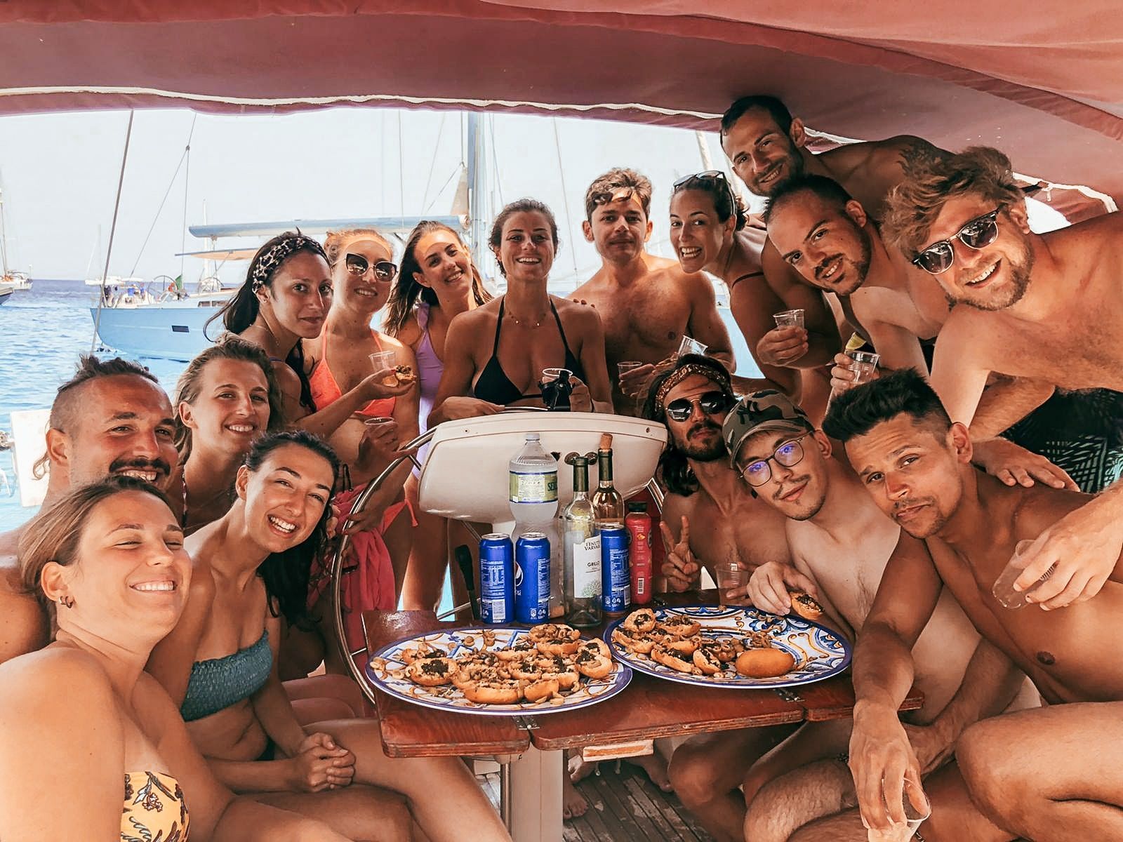 Un gruppo WeRoad si riunisce sul ponte di una barca, sorridendo insieme davanti a cibo e bevande con il mare alle spalle.