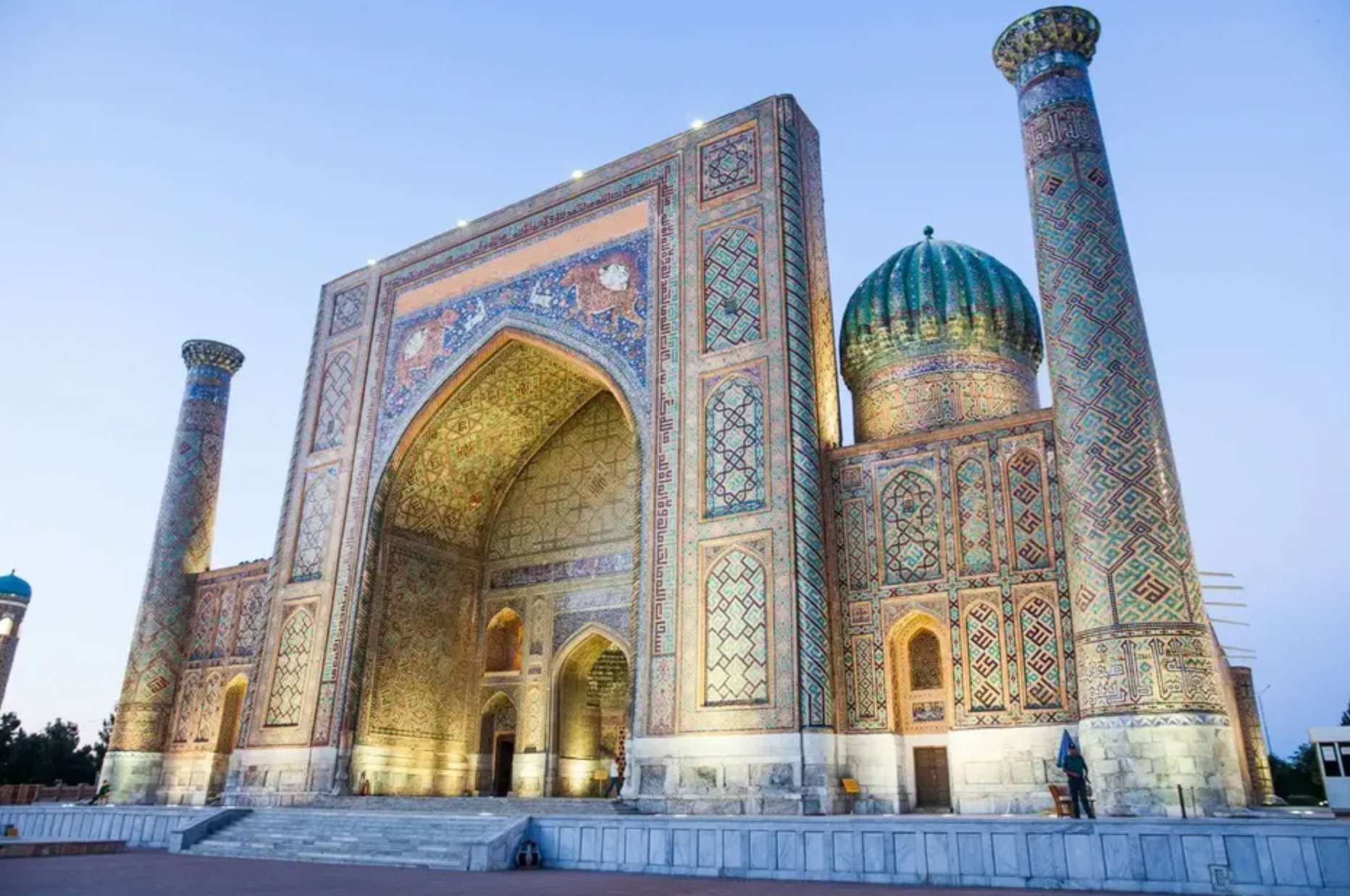 Un edificio riccamente decorato con un grande arco, una cupola turchese e minareti ricoperti di intricati mosaici blu e oro al crepuscolo.
