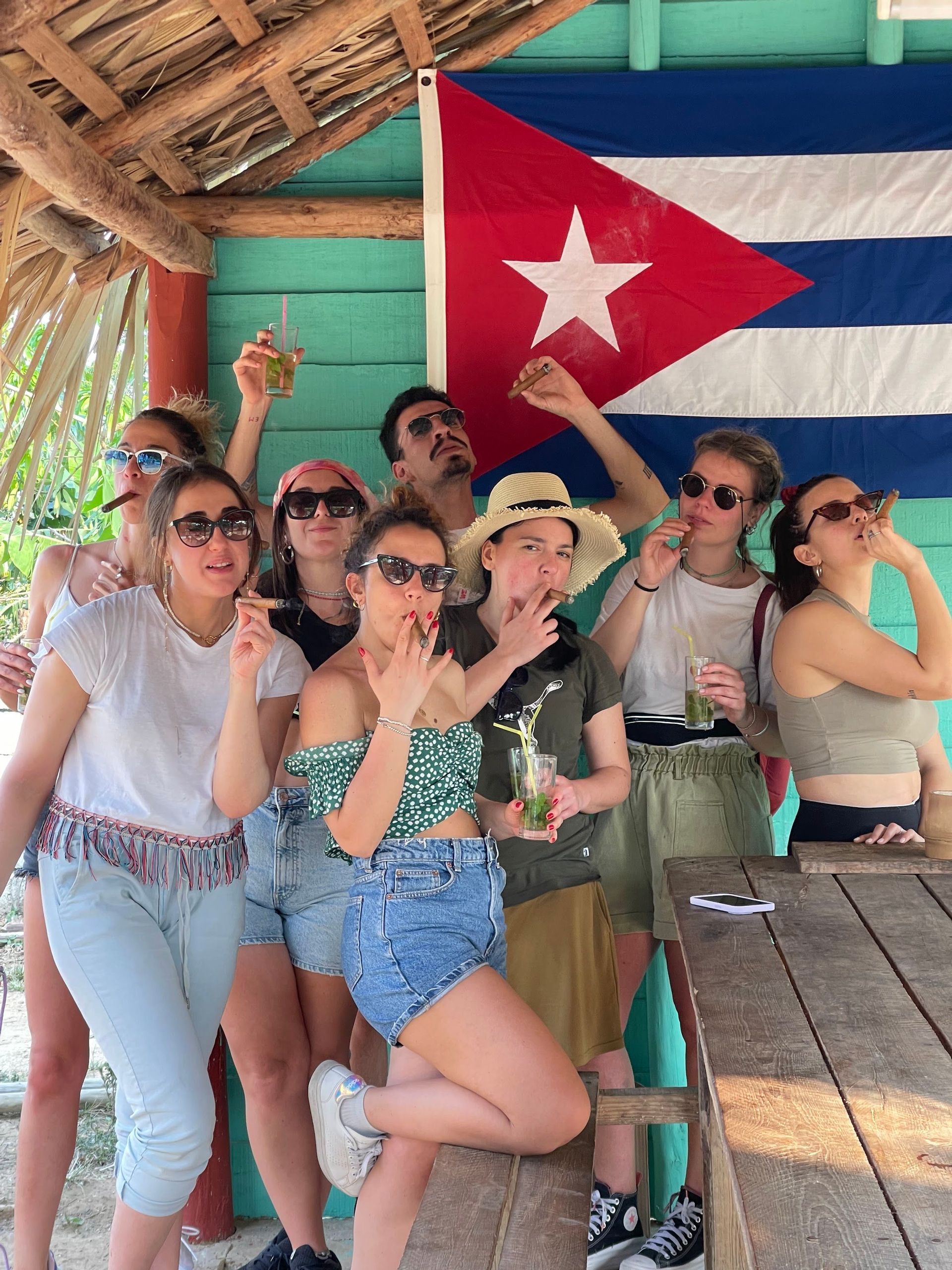 Un grupo de WeRoad posa para una foto fumando puros y bebiendo mojitos frente a una gran bandera cubana.