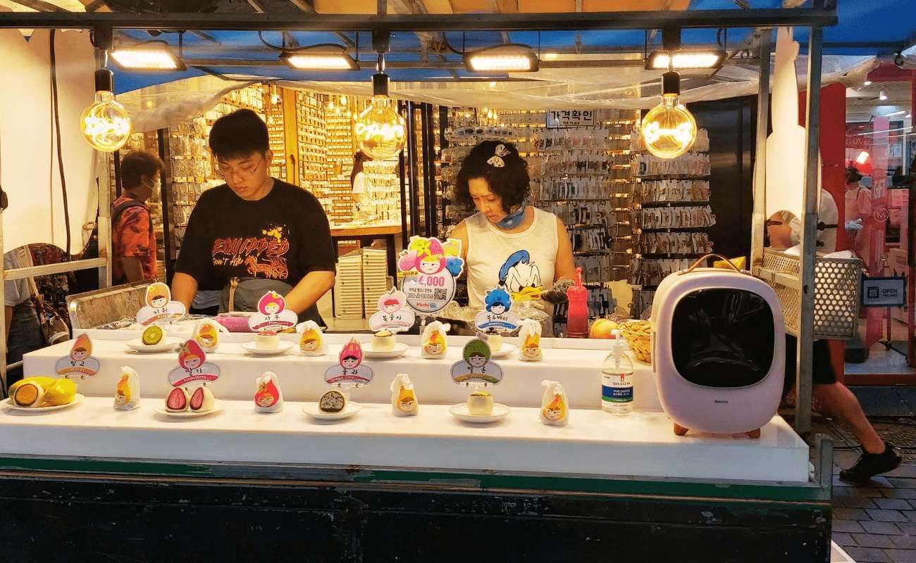 Un uomo e una donna lavorano di notte a una bancarella di street food, preparando piccoli dolci di frutta esposti su un bancone bianco sotto luci calde.