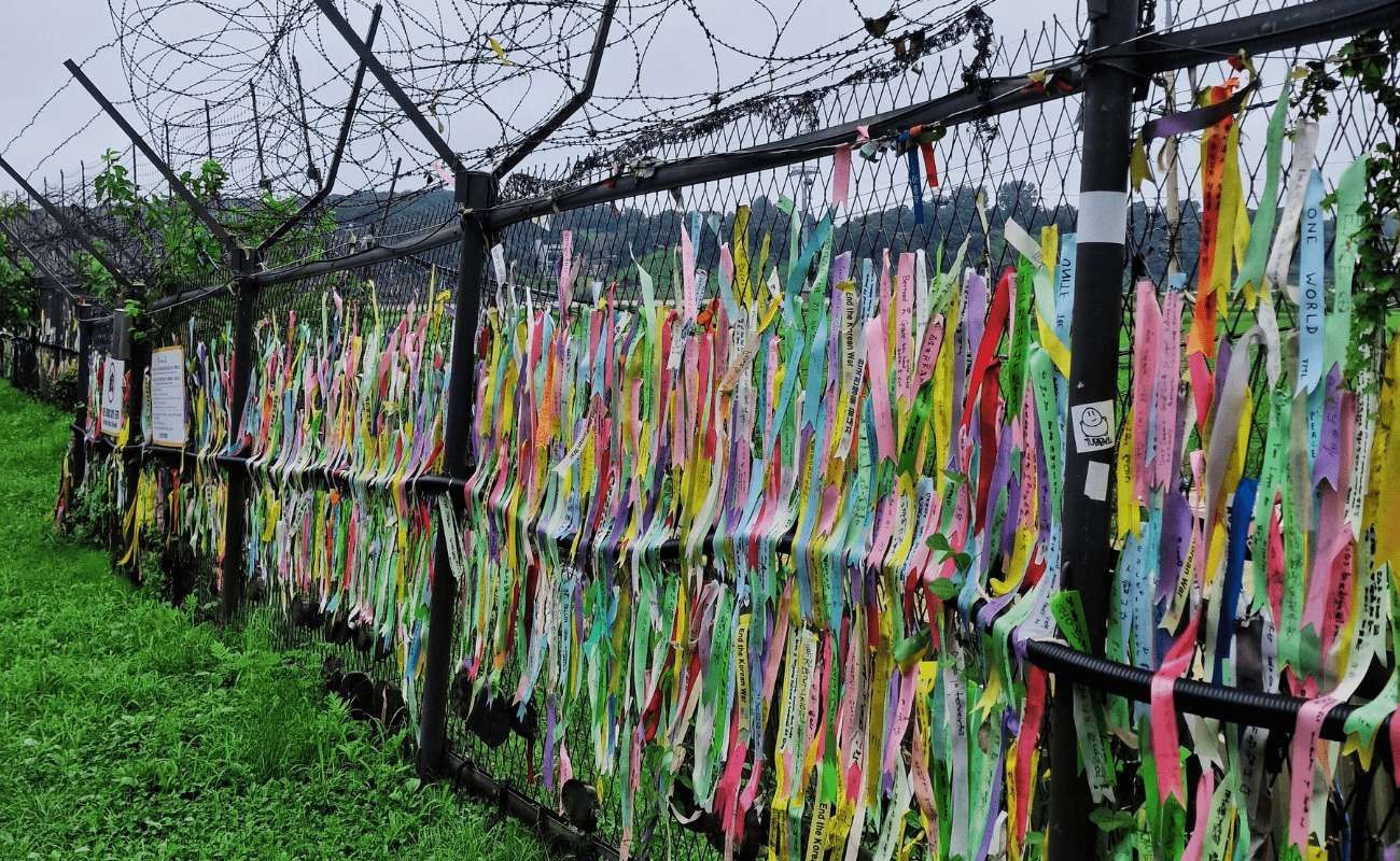 Centinaia di nastri colorati con messaggi scritti sono legati a una recinzione a rete metallica sormontata da filo spinato.