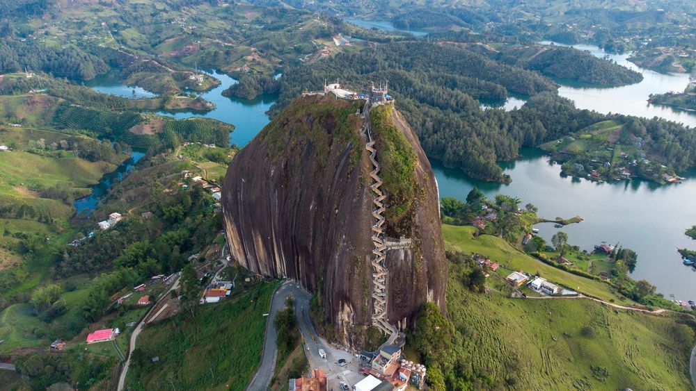 Veduta aerea di un enorme monolito con una scalinata incorporata nel suo fianco, immerso in un paesaggio di verdi colline e laghi blu serpeggianti.