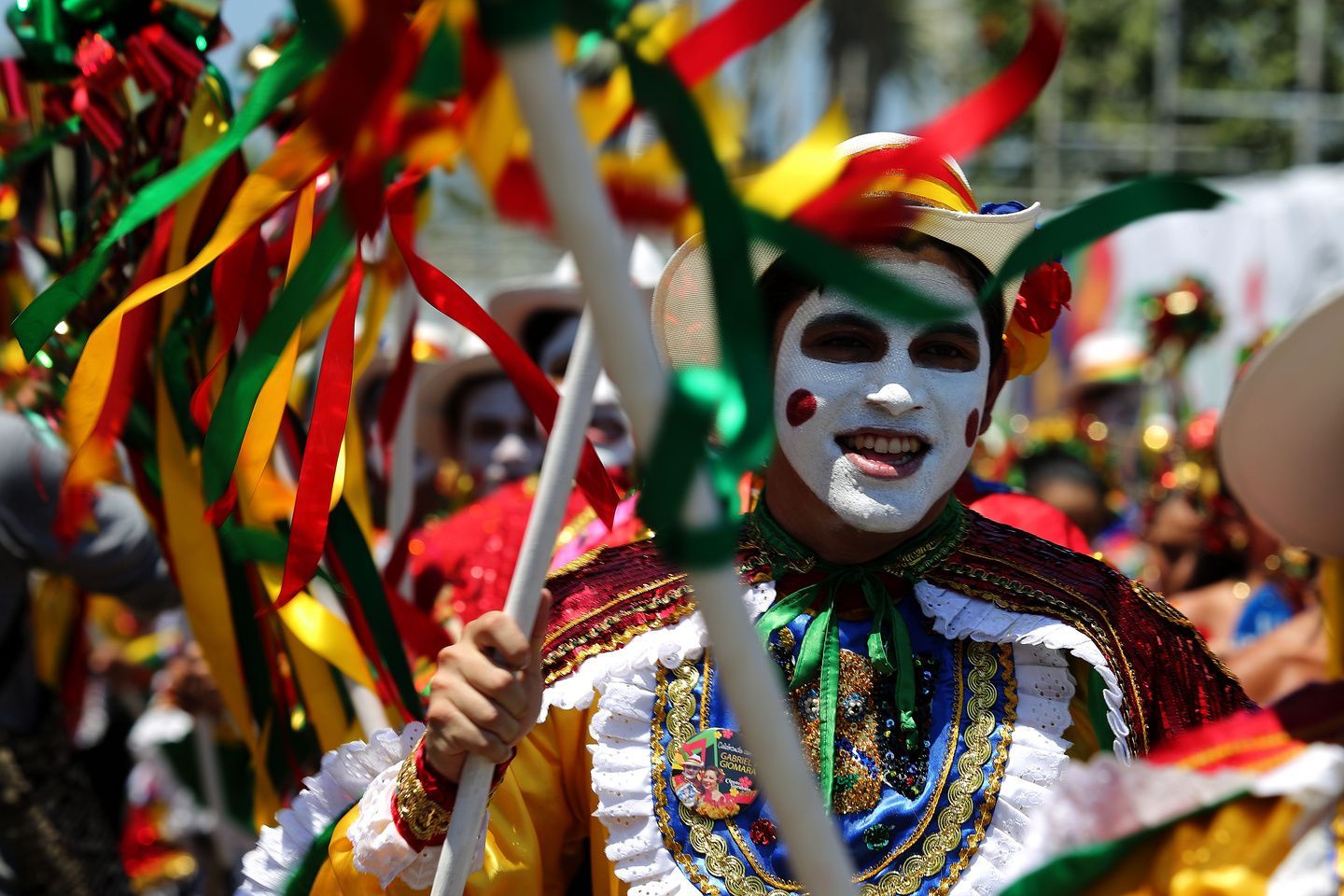Una persona in costume colorato e con il volto dipinto di bianco sorride durante una parata, tenendo un bastone decorato con nastri.