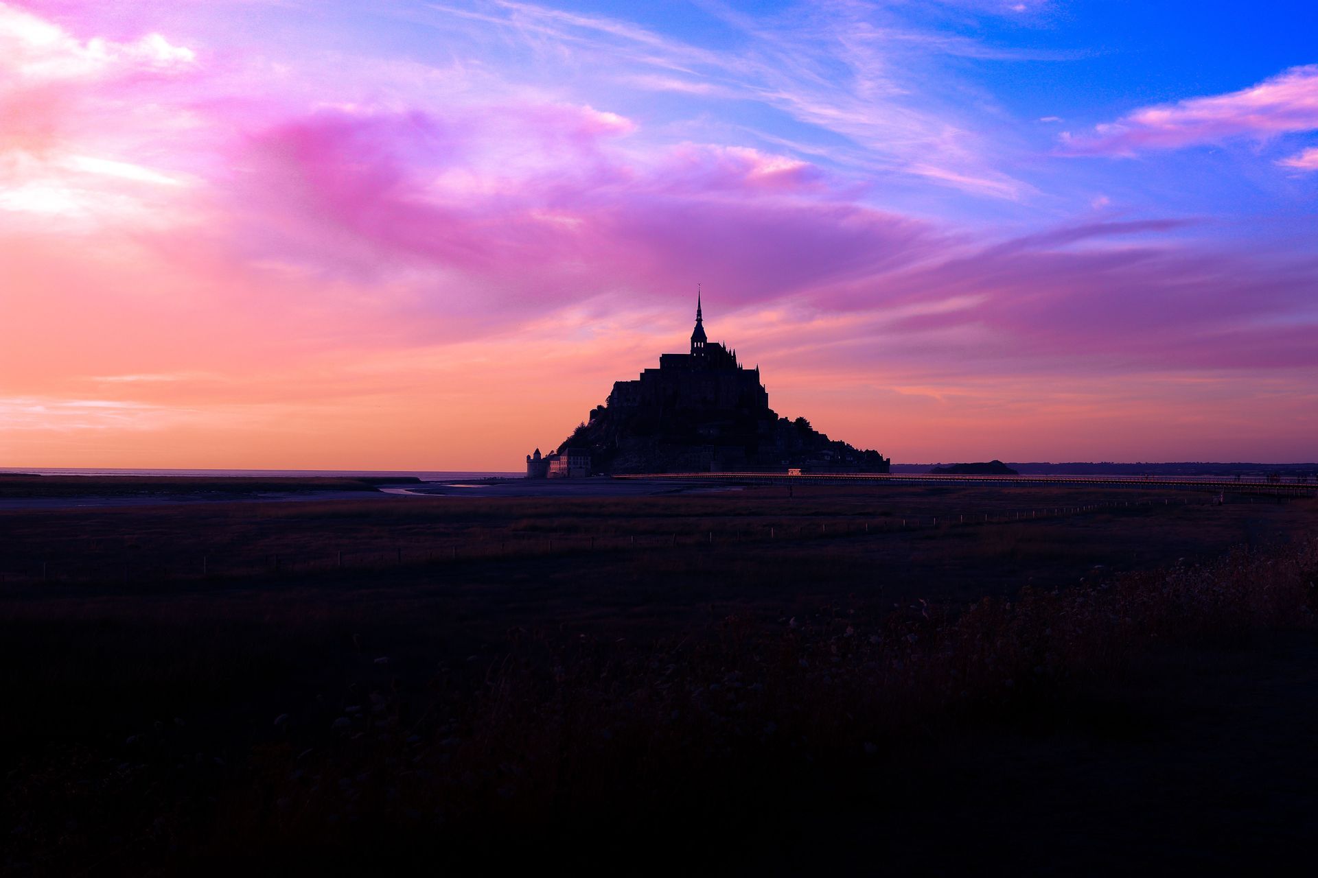 Un château sur une île se silhouettant sur un ciel éclatant avec des nuages roses et violets au coucher du soleil.