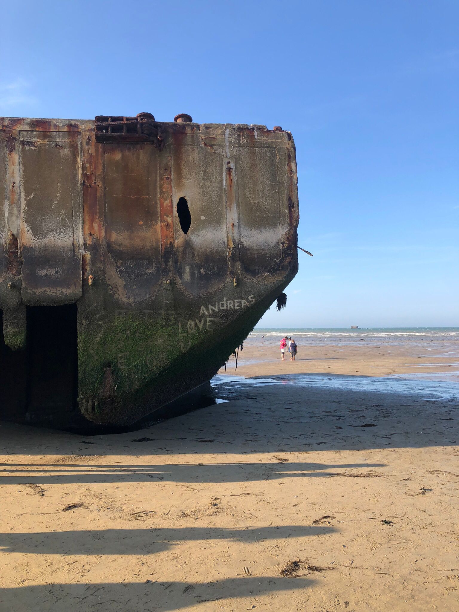 Ein großer, verwitterter Betonblock eines Schiffswracks liegt an einem Sandstrand, und eine Person lugt darüber.