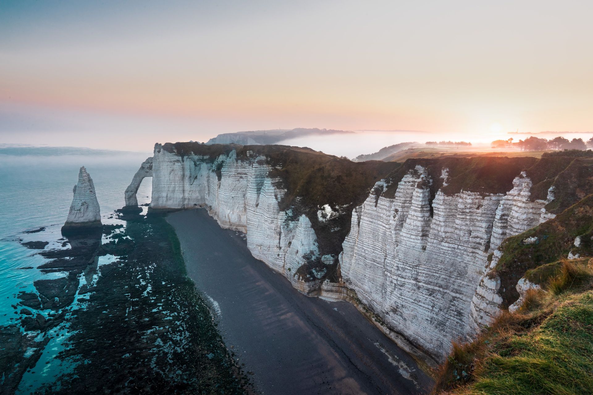 Acantilados de tiza blanca, incluyendo un arco natural y un farallón, se elevan sobre una playa oscura y un mar en calma al amanecer.
