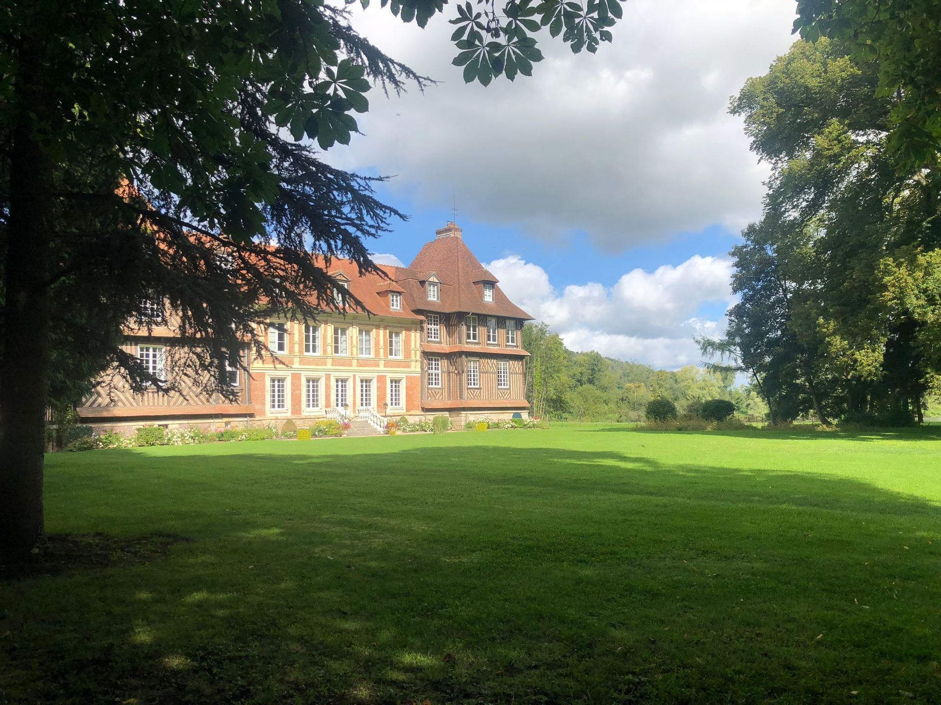 Un grand manoir à colombages avec un toit de tuiles rouges se dresse à côté d&#39;une vaste pelouse verte, encadrée par les branches d&#39;arbres feuillus.