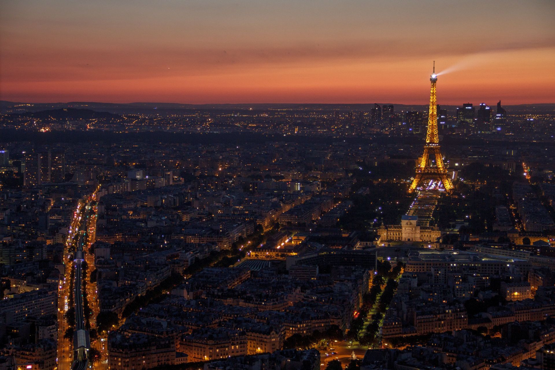 Vue aérienne d&#39;un paysage urbain au crépuscule, avec la Tour Eiffel illuminée projetant un faisceau de lumière sur les lumières de la ville.
