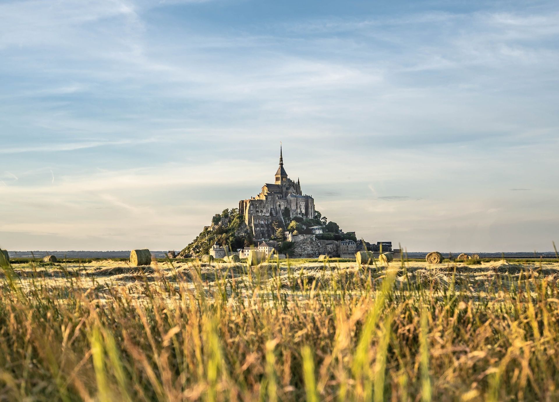 Un castillo histórico en una colina rocosa visto desde un campo de hierba alta y pacas de heno al atardecer.