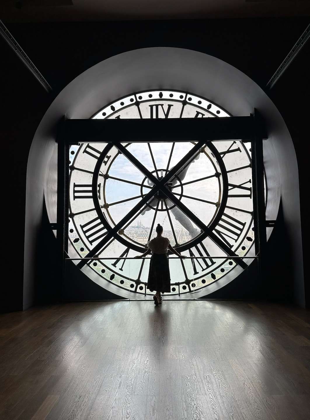Una donna in silhouette si trova su un pavimento di legno, guardando un panorama urbano attraverso una grande finestra a orologio.