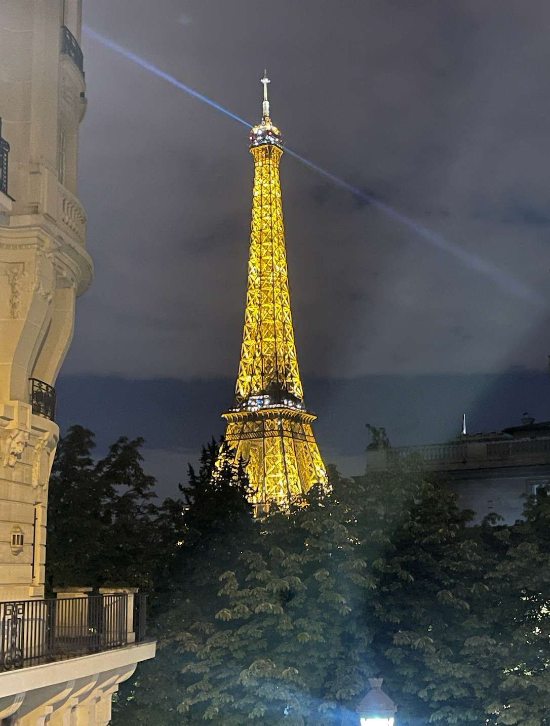 La Torre Eiffel illuminata da luci dorate di notte, il suo faro che proietta un fascio nel cielo nuvoloso, visibile sopra le cime degli alberi.
