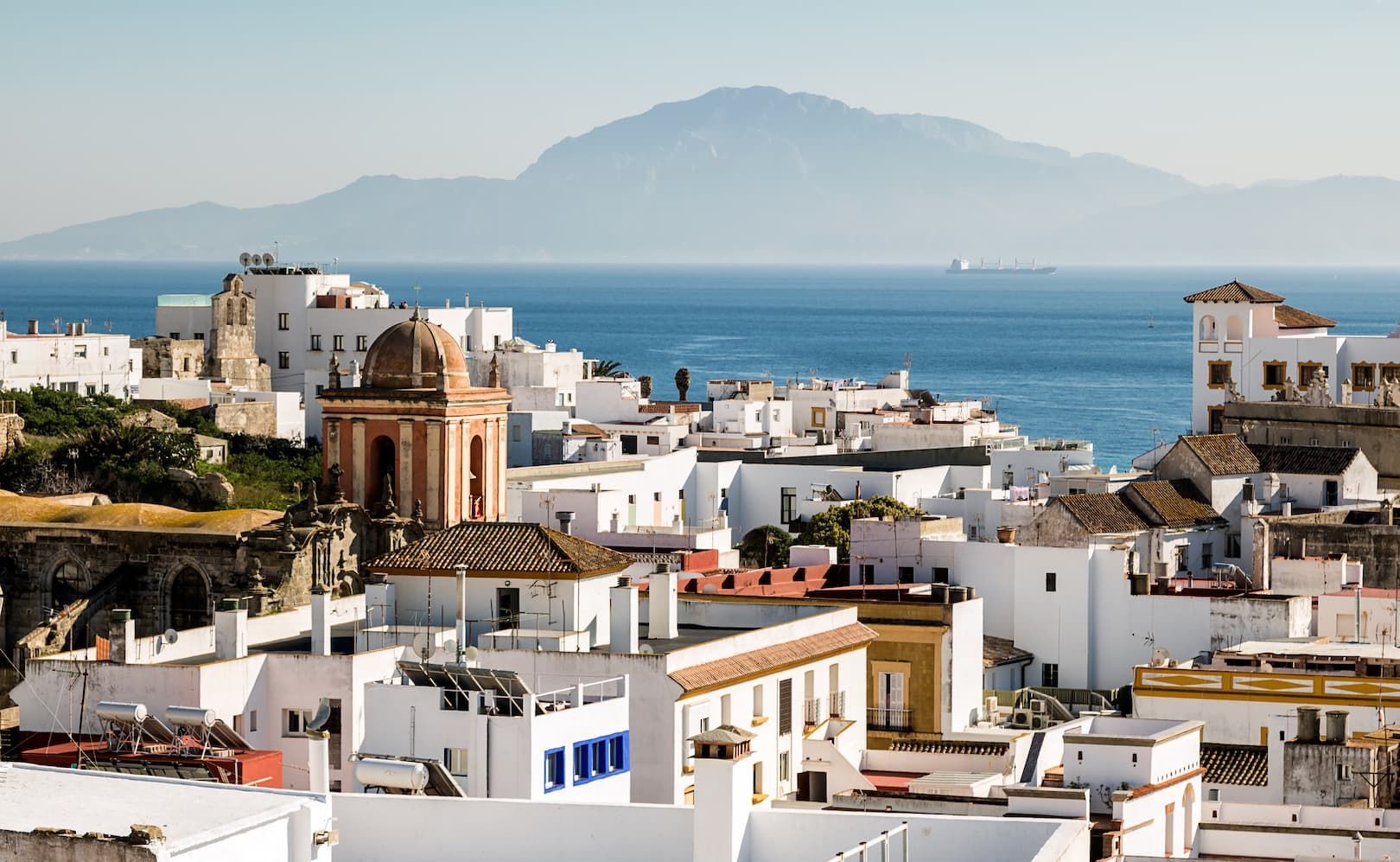 Una vista dall'alto di una cittadina costiera con edifici imbiancati affacciati sul mare e una grande montagna in lontananza.