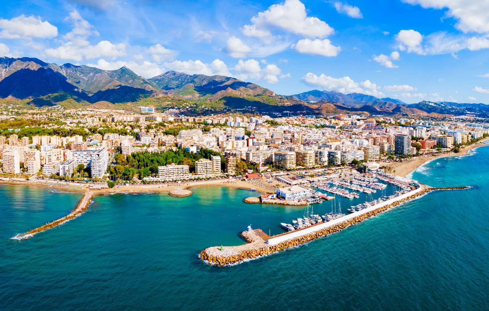 Vista aerea di una città costiera con un grande porto turistico, spiaggia sabbiosa e montagne sullo sfondo sotto un cielo blu e nuvoloso.