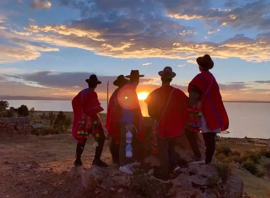 A WeRoad group trip wearing red ponchos and hats watches the sunset over a body of water from a rocky hill.
