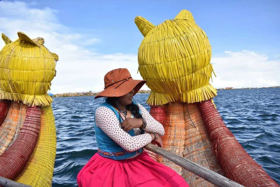 Una donna in abiti tradizionali e un cappello marrone è seduta su una barca decorativa di giunco giallo su un grande specchio d'acqua.