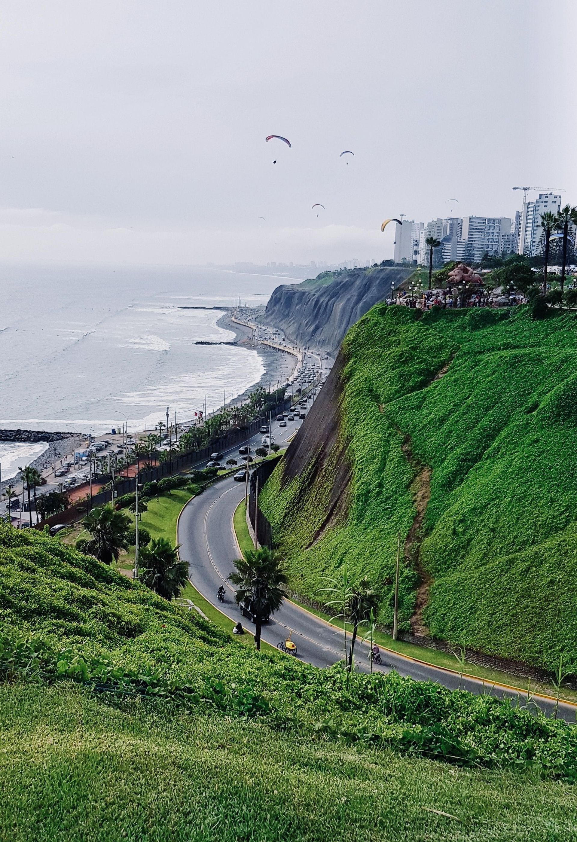 Paraglider gleiten über eine kurvige Küstenstraße und grüne Klippen am Meer, mit Stadtgebäuden im Hintergrund.