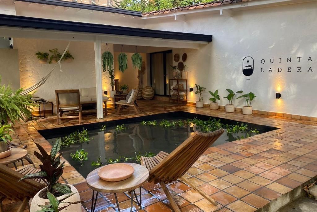 A tiled patio courtyard with a central pond, wooden lounge chairs, a hammock, and many potted plants under warm lighting.