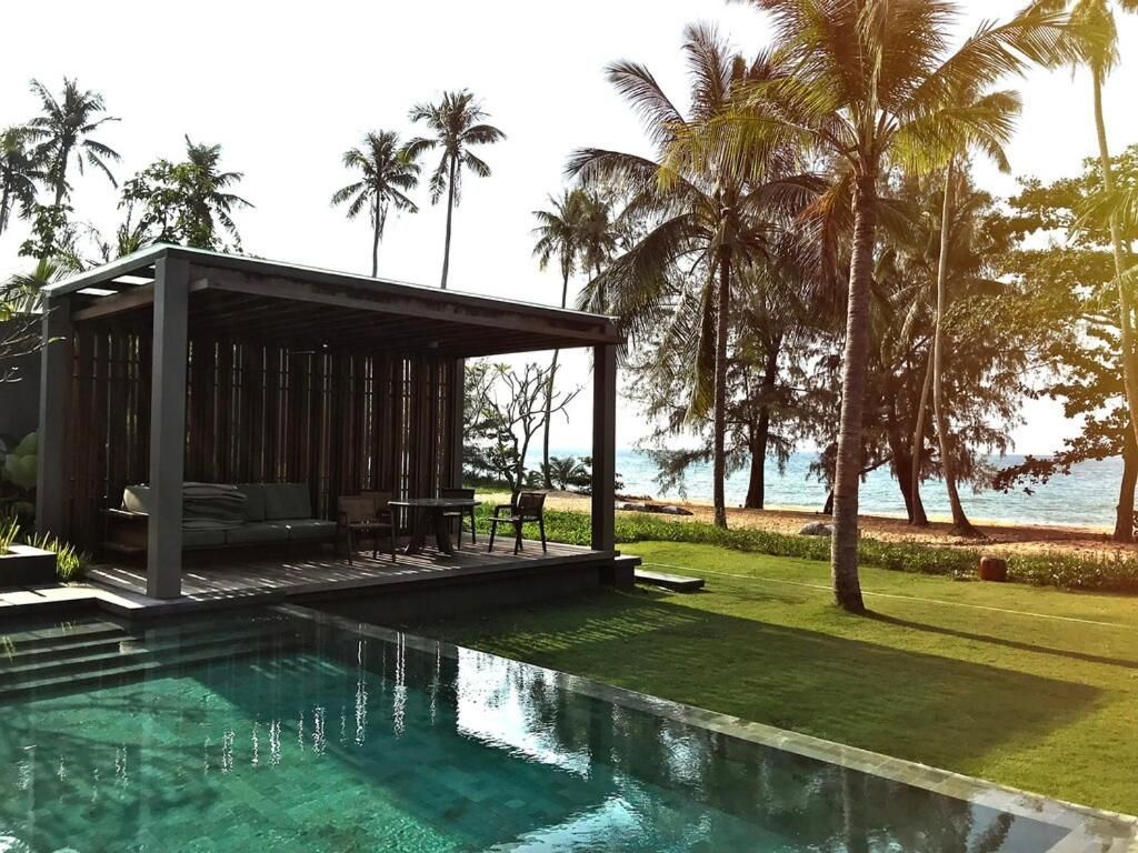 Una piscina e una cabana in legno su un prato erboso, accanto a una spiaggia sabbiosa con palme e l'oceano.