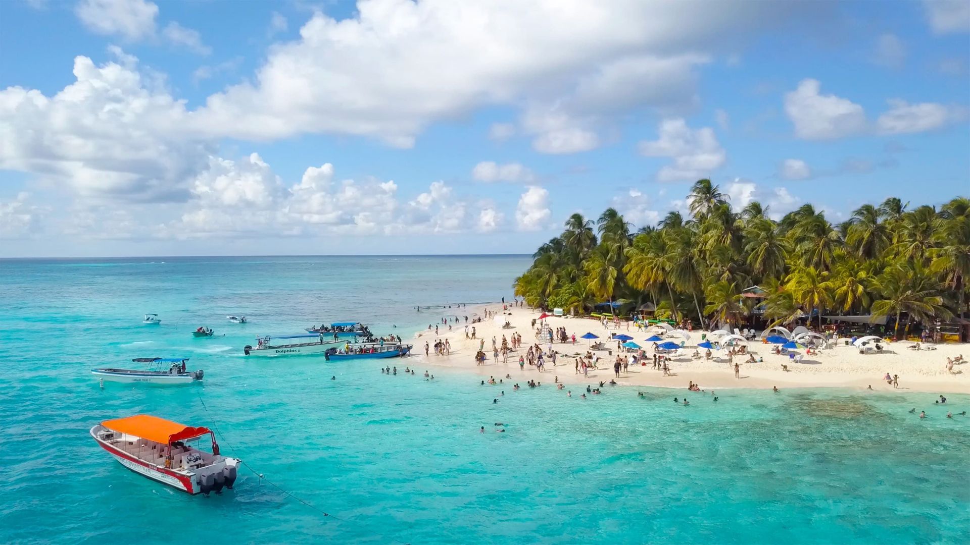 Una vista aerea di una spiaggia tropicale affollata con acqua turchese, numerose imbarcazioni e persone che nuotano e prendono il sole sulla sabbia.