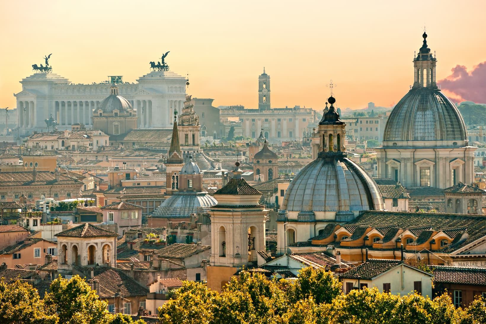A cityscape of historic domes, monuments, and buildings with terracotta roofs seen from above under a hazy golden sky.
