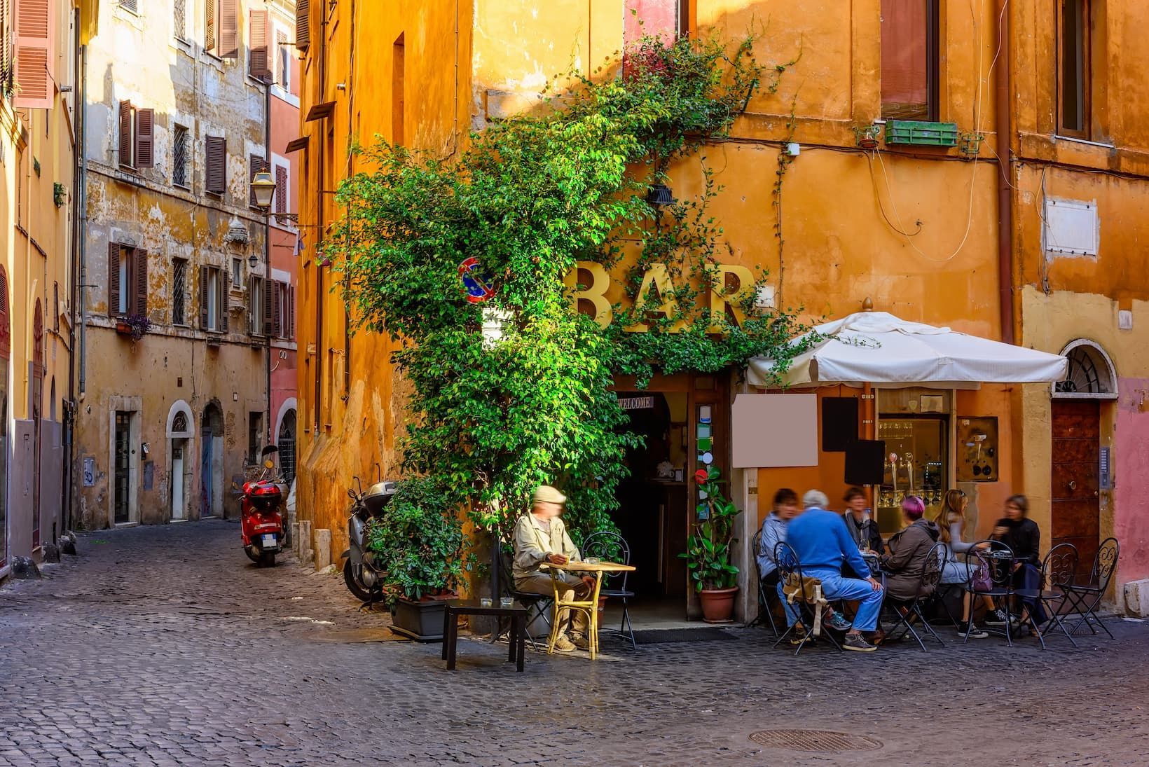 A sunny cobblestone alley with people sitting at an outdoor bar covered in lush green ivy.